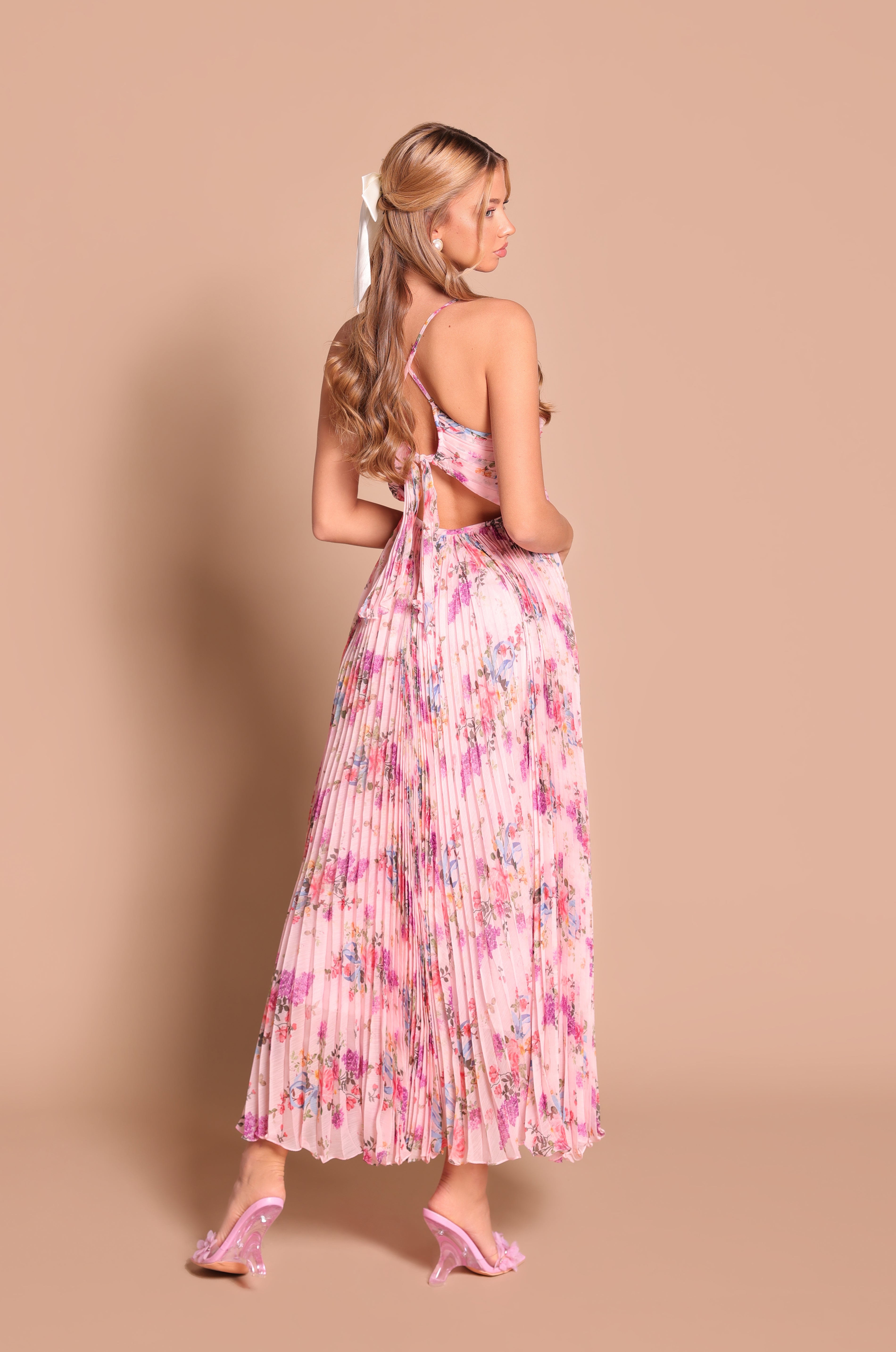 Woman with long hair wears the Pleated Blush Pink Floral Maxi Dress: Florinne and matching heels, standing sideways and facing backward against a beige background.